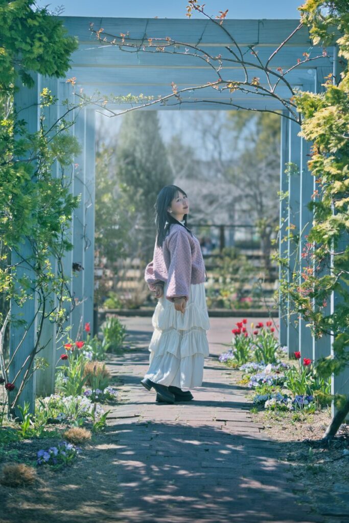 一面に広がる花畑の中、背を向けて立つモデル・ぬぬの広角ポートレート。奥には山並みが連なる、晴天の下での自然な記録。Luminar Neoの「太陽光線」適用前の原版。