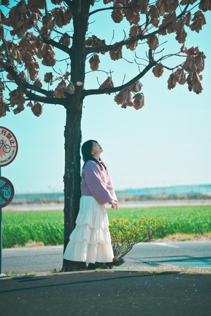 枯葉の残る木の下に立ち、静かに空を見上げるモデル・ぬぬ。24mm f/1.7レンズで捉えた、物語のワンシーンのような情緒あふれる全身構図