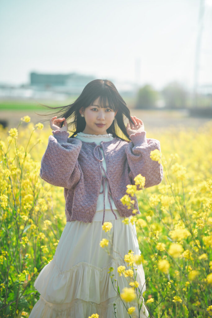 菜の花畑の中で髪に手を添えるモデル・ぬぬ。24mm単焦点レンズの豊かな描写性能と、Luminar Neoによる色彩調整のポテンシャルを示す一枚。