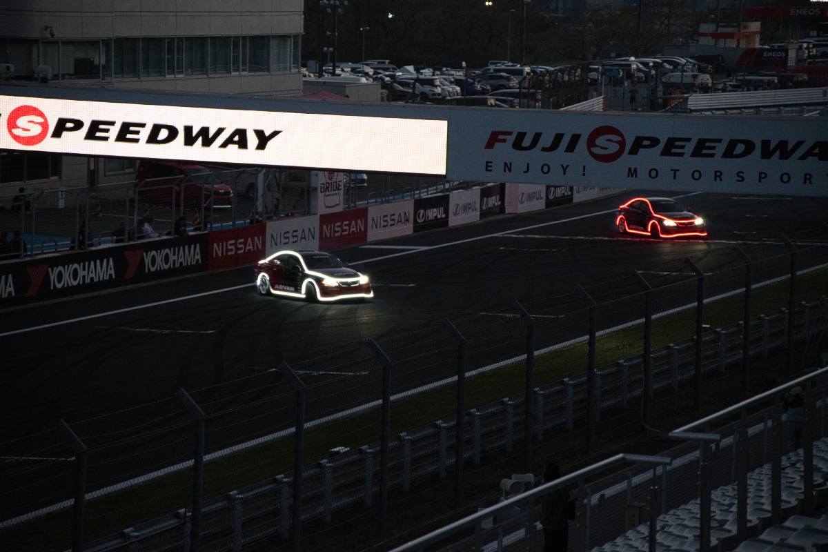 富士スピードウェイの観覧席から、電飾で縁取られた2台のマシンがホームストレートを駆け抜ける様子