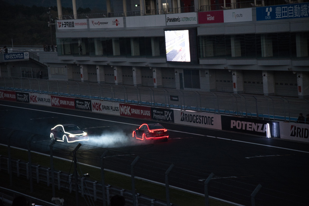 「ADVAN」と「RAYS」のロゴが輝くフェンス越しに、コースを流れるマシンの鮮やかな光跡