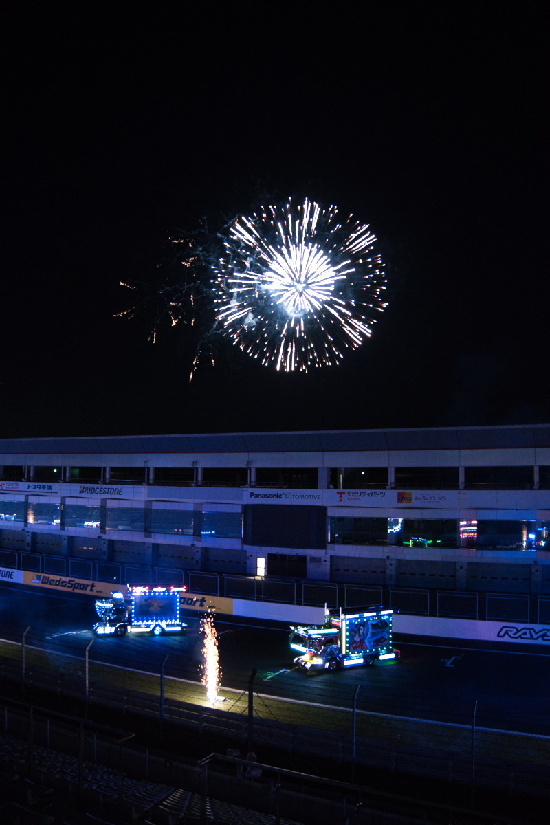 夜のサーキット上空、電飾マシンの頭上で一際大きく、白く輝く単発の打ち上げ花火
