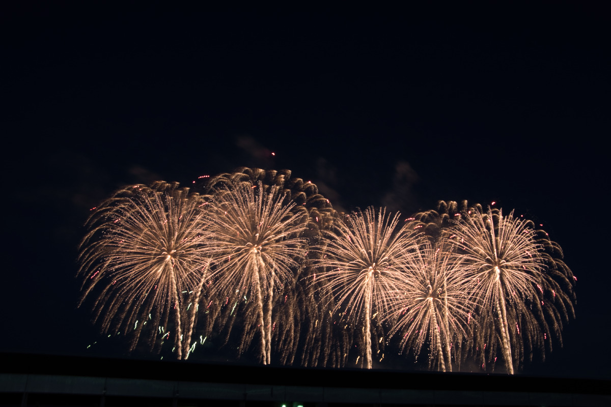 富士スピードウェイの施設越しに、空を埋め尽くすように横並びで咲き誇る四輪の黄金色の花火