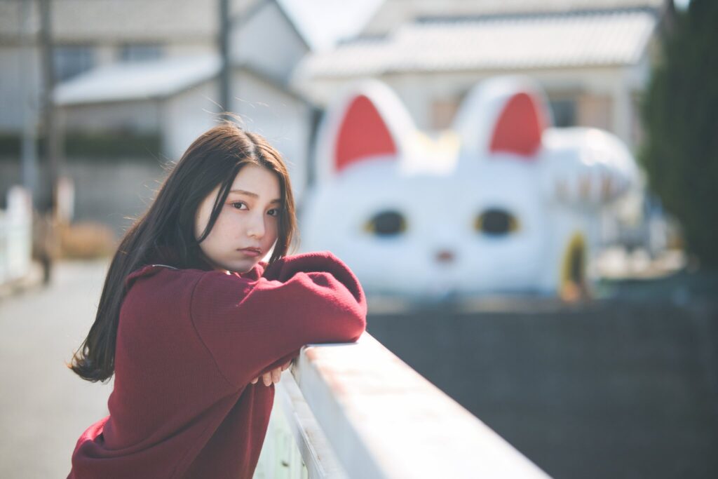 巨大招き猫「とこにゃん」を背景に、欄干に身を乗り出す花咲ゆいなさんのポートレート