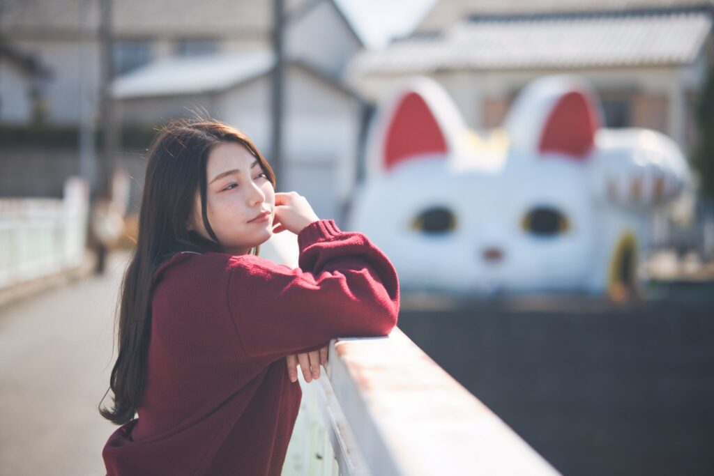 巨大招き猫「とこにゃん」を背景に、欄干に身を乗り出す花咲ゆいなさんのポートレート