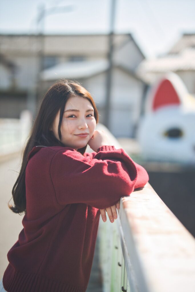 巨大招き猫「とこにゃん」を背景に、欄干に身を乗り出す花咲ゆいなさんのポートレート
