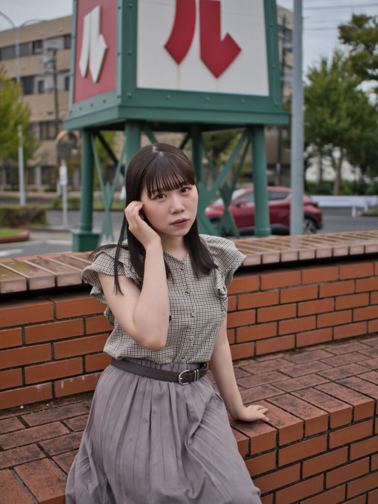カブトビールの看板の前に座るモデル。雨の日の外光を利用し、レンガの質感とゆめのさんの笑顔を捉えたポートレート。LUMIX G100Dの「撮って出し」の底力。
