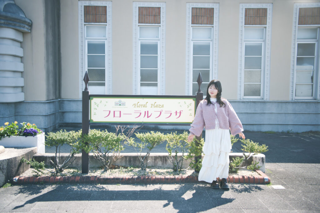 「フローラルプラザ」の看板の横に立ち、両手を広げてポーズをとる女性。背景には洋風の建物が見える。