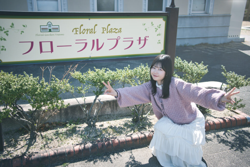 「フローラルプラザ」の看板の前でしゃがみ、両手でピースサインを作る笑顔のぬぬさん。