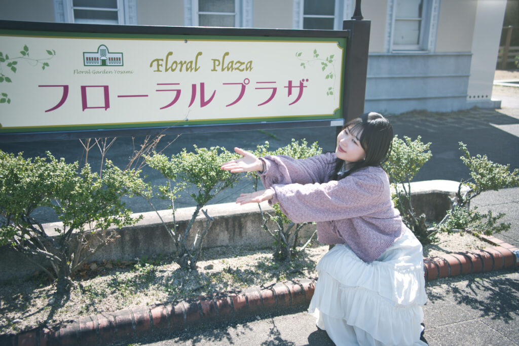 「フローラルプラザ」の看板の前でしゃがみ、斜め上を向いて両手を差し出すポーズをとる女性。