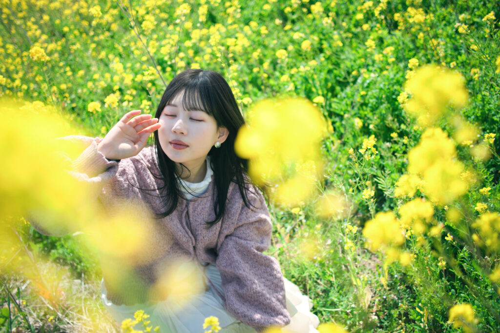 菜の花に囲まれて座り、目を閉じて顔に手を添える女性のクローズアップ。手前の菜の花が柔らかくボケている。