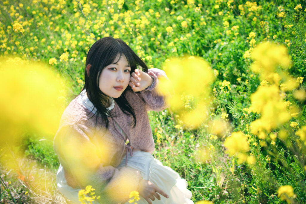 菜の花に囲まれて座り、目を閉じて顔に手を添える女性のクローズアップ。手前の菜の花が柔らかくボケている。