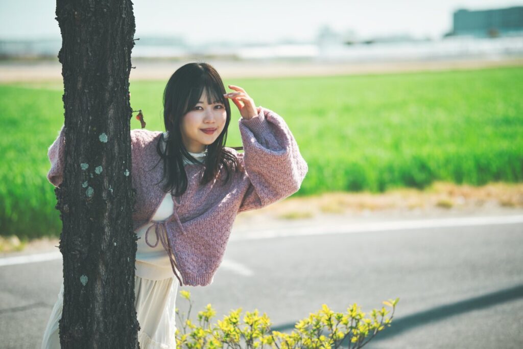 木の後ろからひょっこりと顔を出し、微笑むモデル・ぬぬ。24mm f/1.7のレンズが描き出す背景の緑とボケ味を活かした、奥行きのあるポートレート。