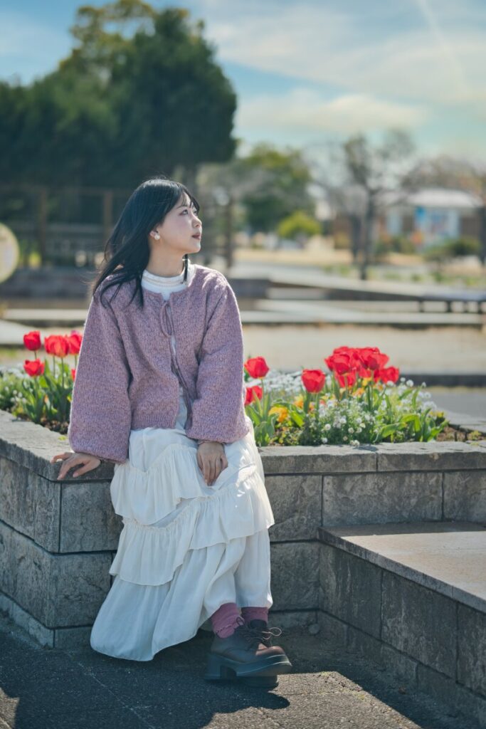 赤いチューリップが咲く公園の縁に座り、遠くを見つめるモデル・ぬぬの全身ポートレート。Luminar Neoによる空の再構築とカラー調整で、あの日感じた春の空気感を表現した「記憶」としての作品。