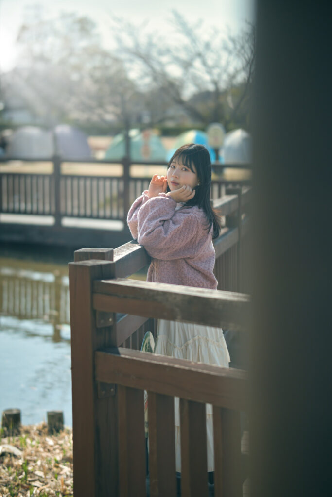 公園の木の橋の欄干に身を乗り出し、柔らかな光の中でこちらを見つめるモデル・ぬぬのポートレート。手前の柱を前ボケに利用した奥行きのある構図。Luminar Neo適用前の原版。