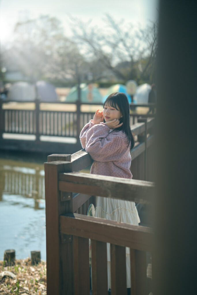 公園の木の橋の欄干に身を乗り出し、柔らかな光の中でこちらを見つめるモデル・ぬぬのポートレート。手前の柱を前ボケに利用した奥行きのある構図。Luminar Neo適用前の原版。