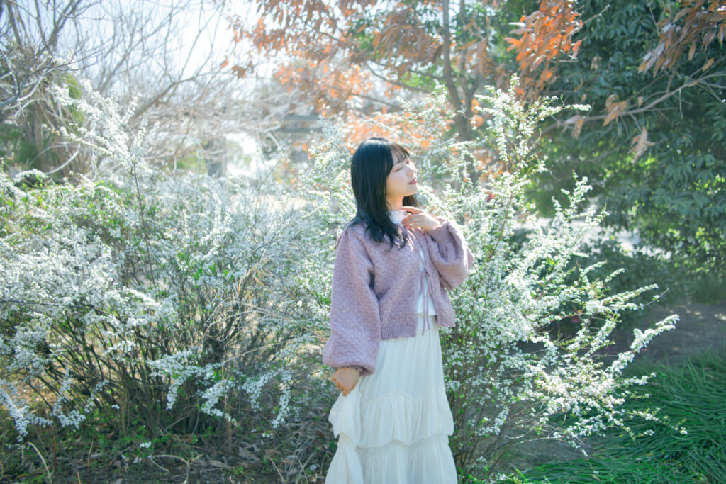 花に囲まれて座り、目を閉じて顔に手を添えるぬぬさん。手前の菜の花が柔らかくボケている。