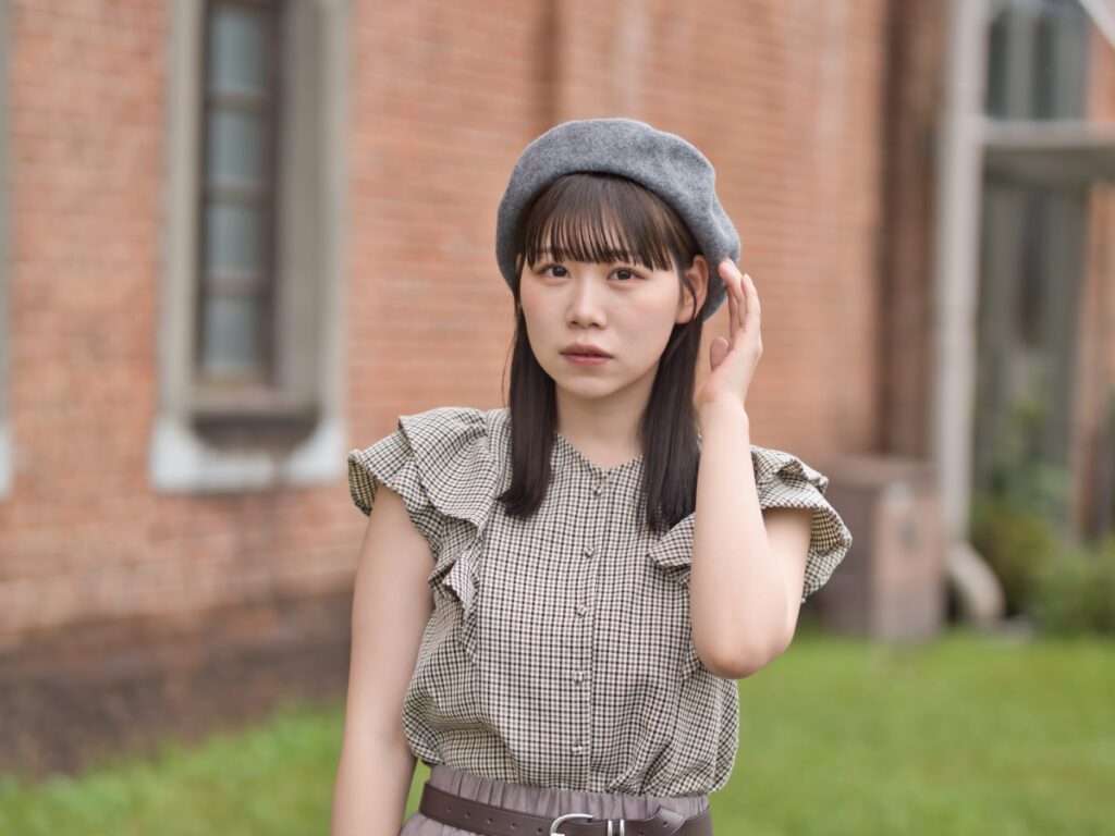 赤レンガの壁の前で、自然な笑顔を見せるゆめのさんのポートレート。F1.4の絞り開放による、被写体を浮かび上がらせる柔らかな背景ボケ。