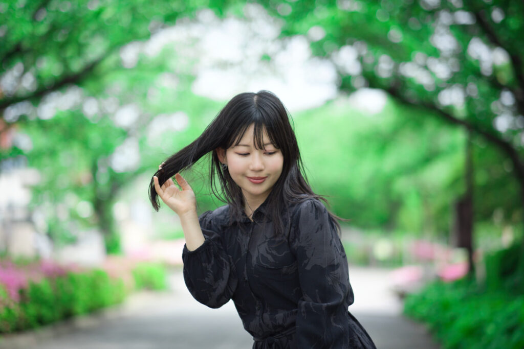 新緑の並木道でXF56mmF1.2 Rを使用し、光が優しく回り込む中で髪を整えるぬぬさんのナチュラルなポートレート。