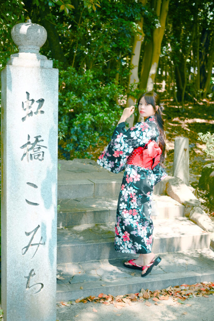 「虹橋こみち」の石柱の横で、神社の緑を背景に佇む海月みゆきさんの全身像