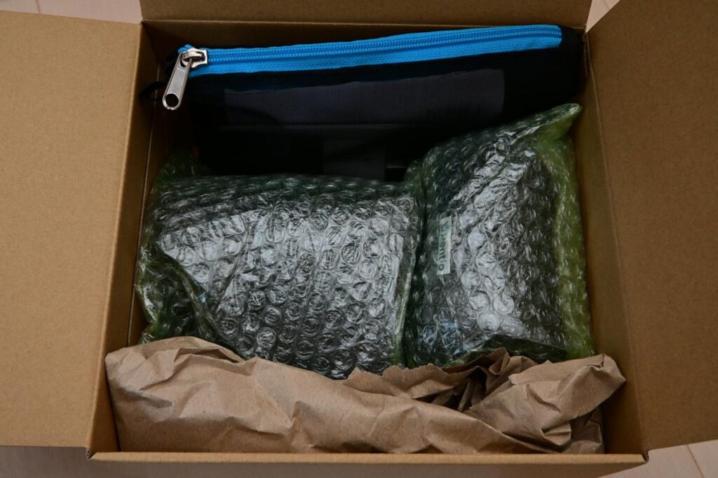 緑色の緩衝材（プチプチ）に包まれたカメラ機材と、丁寧に配置された書類一式が入った段ボール箱の内部