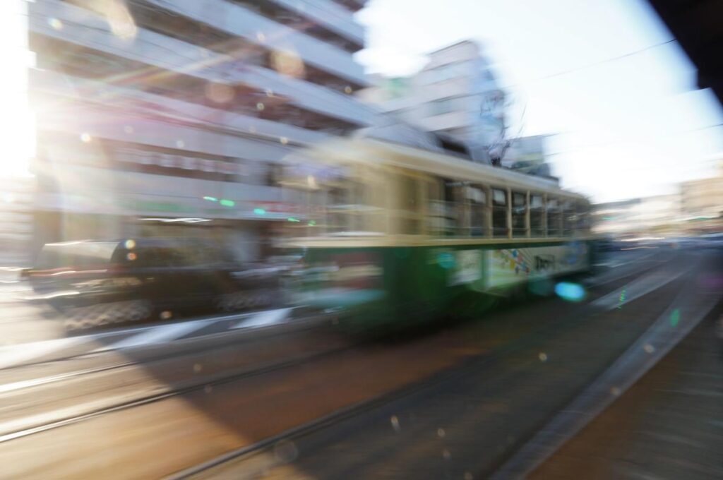 ソニー NEX-F3の流し撮りで撮影された、昼間の街中を走行する緑色の路面電車