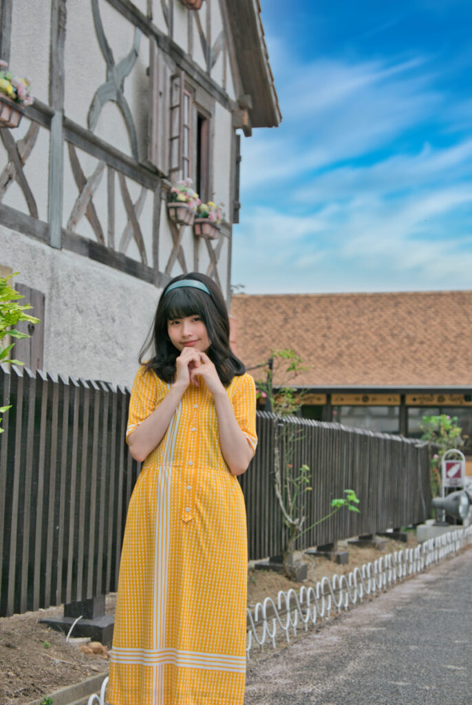木組みの壁と花が飾られた窓辺を背景に、顎の下で手を組む梨乃さんのバストアップ写真