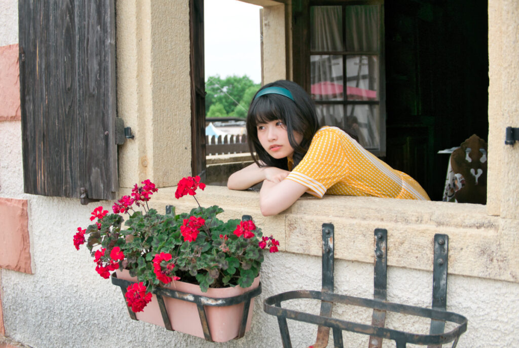 赤い花が咲く窓辺に両肘を置き、アンニュイな表情でこちらを見つめる梨乃さんの写真