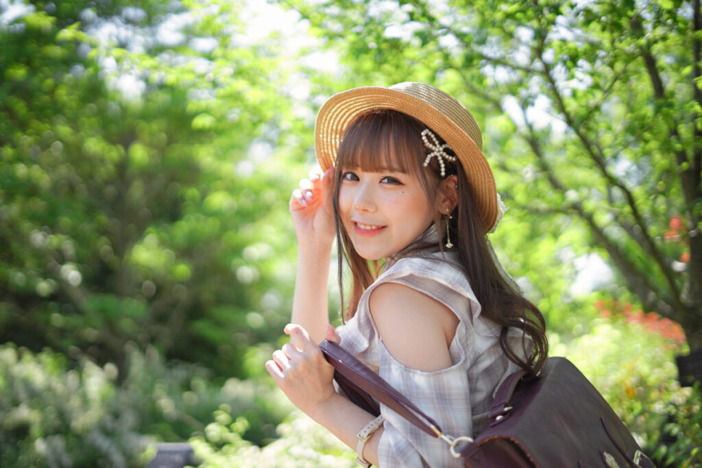 背景の新緑が綺麗な円形の玉ボケとなり、麦わら帽子を押さえながら振り返って弾けるような笑顔を見せる鹿乃ばんびさん。