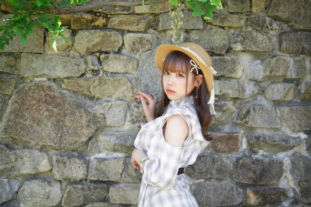 風情のある石壁の前に立ち、少し物憂げな表情で横を向いて佇む鹿乃ばんびさんの上半身カット。