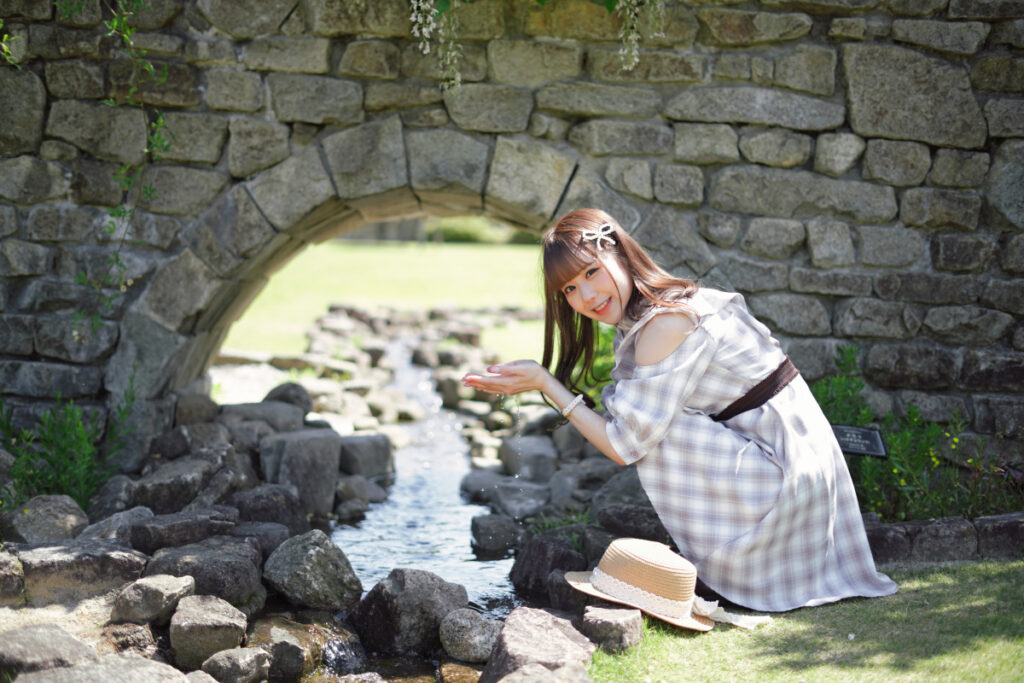 石のトンネルの下を流れる小川のほとりでしゃがみ込み、両手で水をすくうようなポーズで笑顔を見せる鹿乃ばんびさん。