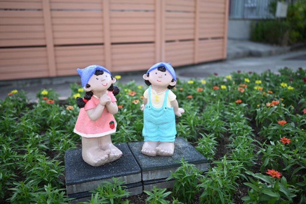 開放f1.8の滑らかな背景ボケを活かして花壇の中に佇む男の子と女の子の人形を捉えたカット