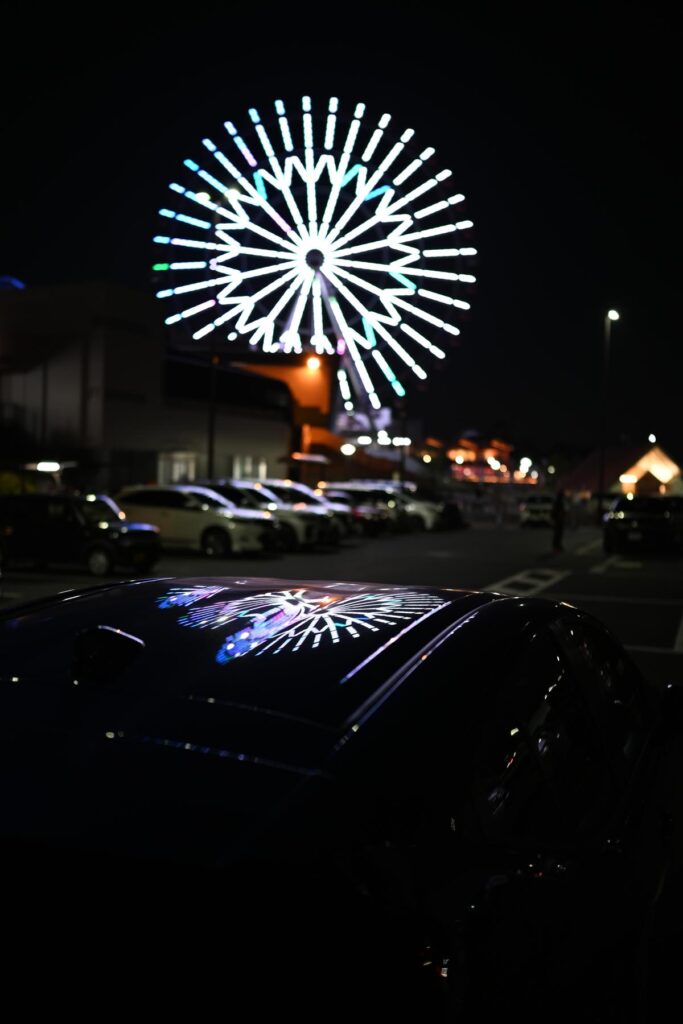 車のルーフに反射する大観覧車のイルミネーションにピントを合わせ背景を大きくぼかした夜景スナップ