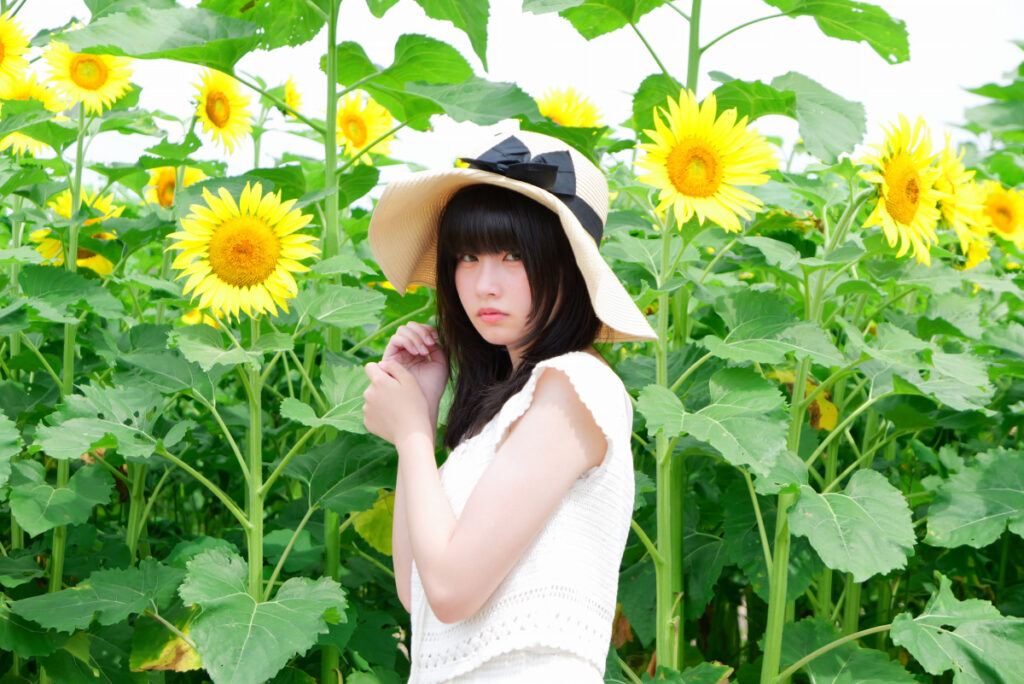 75mm望遠端:物語を感じさせる一本ひまわり前で佇む女性モデルのポートレート