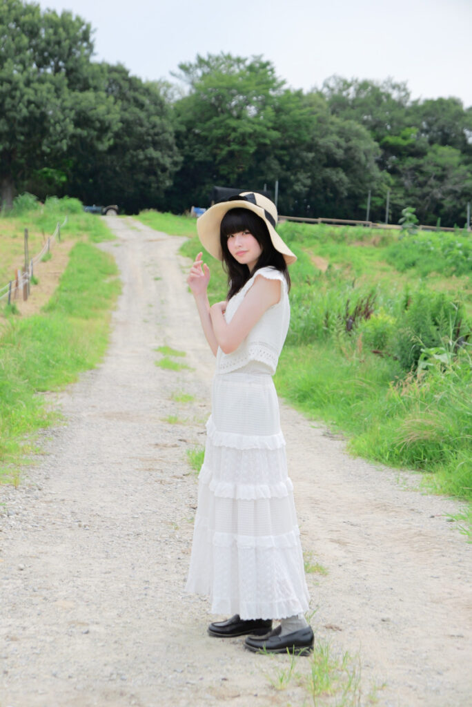 75mm望遠端:まっすぐ続く牧場の一本道を背景に立つ女性モデルの全身ポートレート