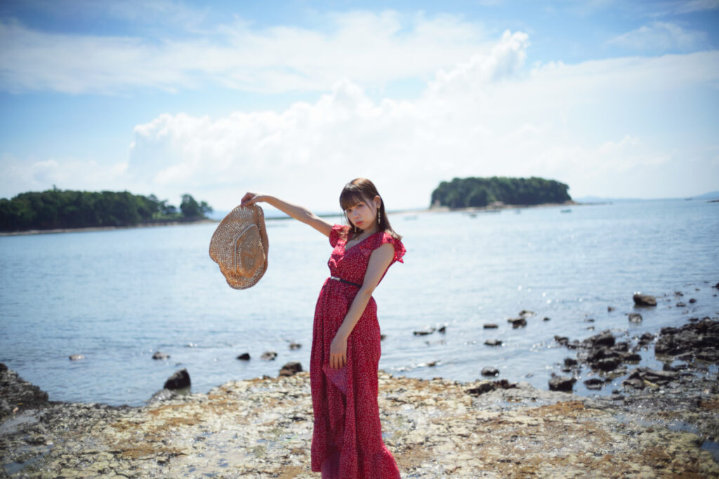 海岸沿いで麦わら帽子を大きく広げ、ポーズをとる鹿乃ばんびさんの引き構図カット