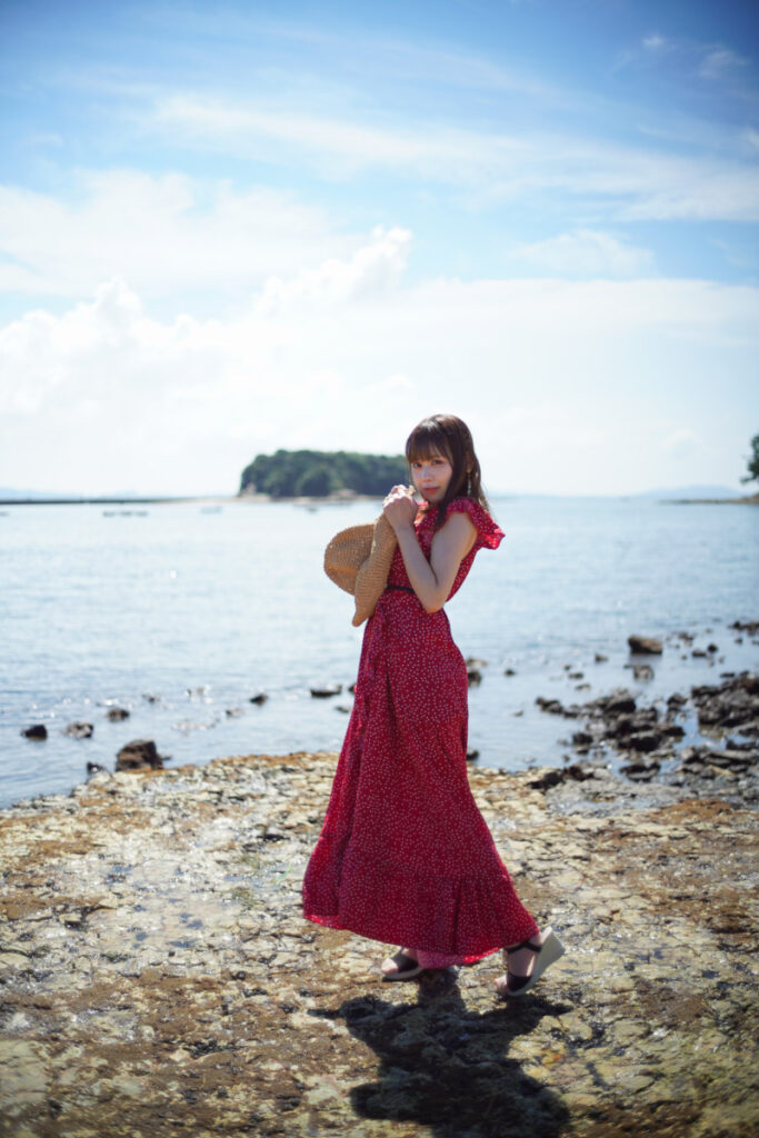 海岸沿いの美しい景色の中で、麦わら帽子を胸に抱いて振り返る鹿乃ばんびさんのポートレート