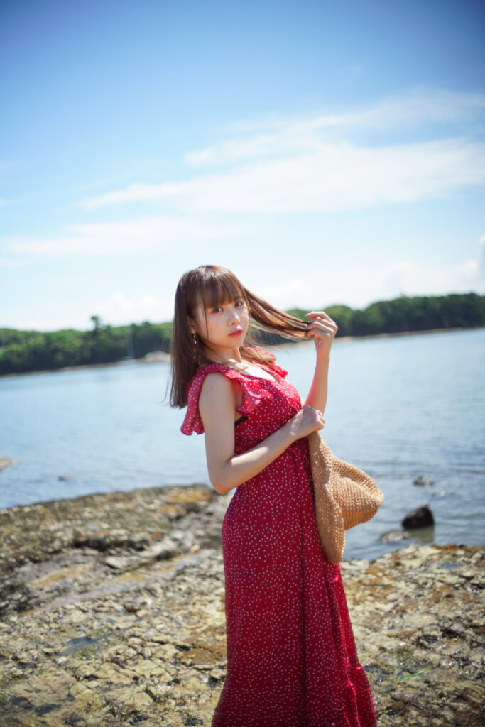 穏やかな海と夏の青空を背景に、麦わら帽子を持って振り返る赤いワンピース姿の鹿乃ばんびさんの印象的なポートレート
