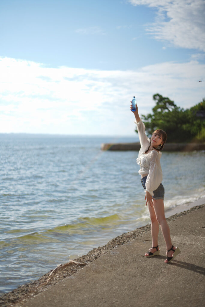 夏の青空と海を背景に、堤防の上で青いボトルを高く掲げて笑顔を見せる鹿乃ばんびさん