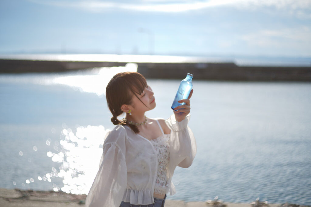 水面のキラキラとした美しい玉ボケを背景に、青いボトルを顔の横に添える鹿乃ばんびさん