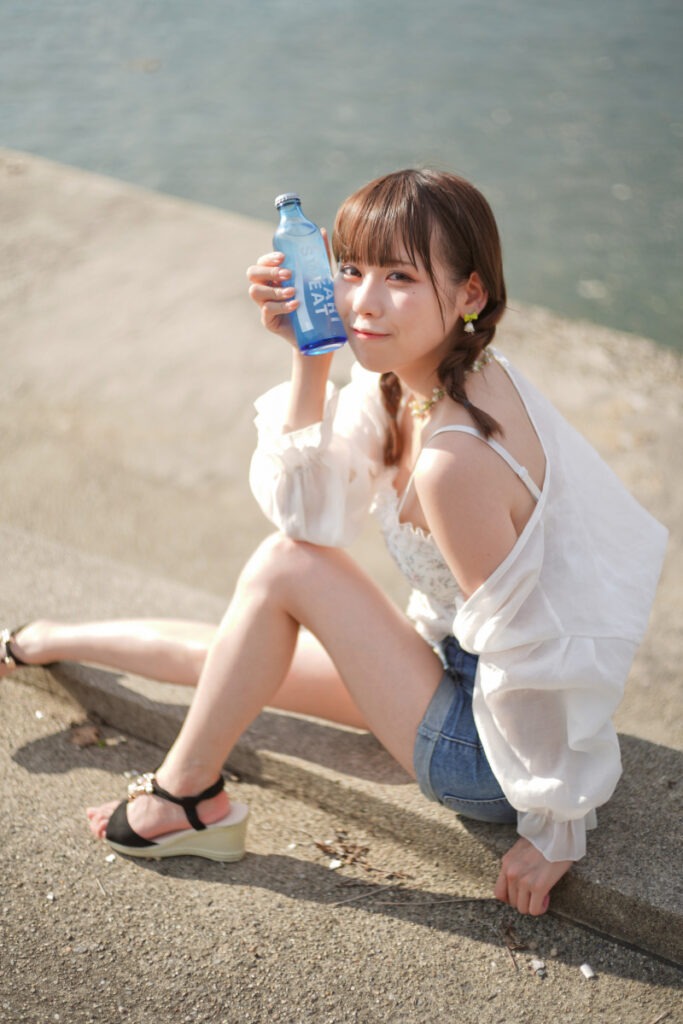 堤防の上で佇み、両手で持った青いボトルをじっと見つめる鹿乃ばんびさんの横顔カット