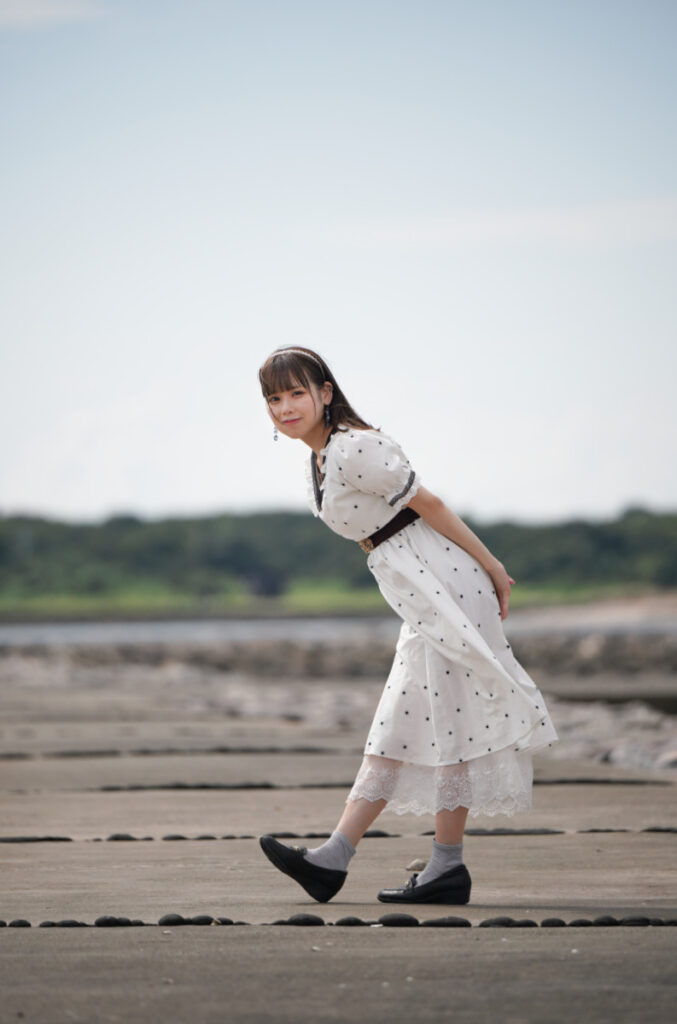 堤防の上でカメラに向かってひょっこりと体を傾ける、白いワンピース姿の鹿乃ばんびさんの180mmカット