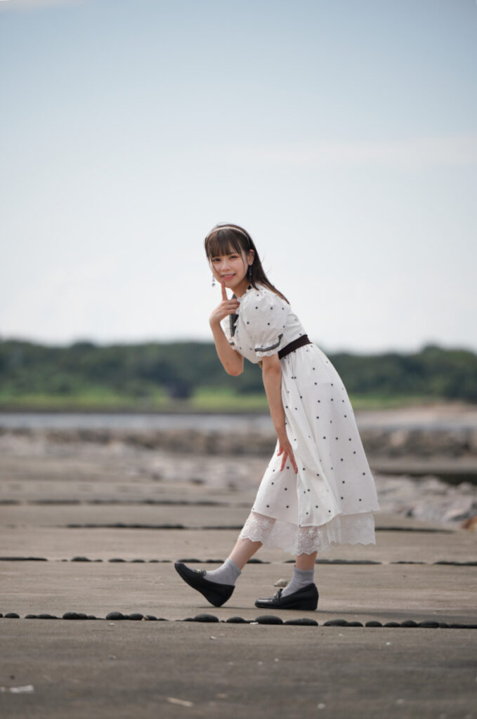 離れた位置から180mmの開放F2.8で捉えた、堤防の上で人差し指を口元に当てる鹿乃ばんびさん