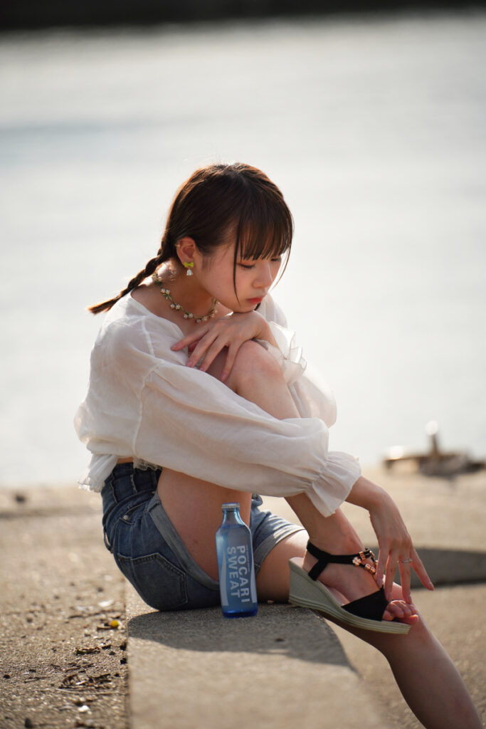 光る水面をバックに、堤防の上で膝を抱えてアンニュイな表情を見せる鹿乃ばんびさんの座りポートレート