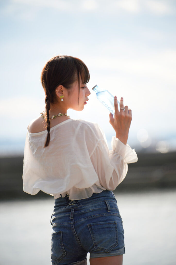 夕暮れの逆光を受けながら、青いボトルを手にして振り返る鹿乃ばんびさんの後ろ姿