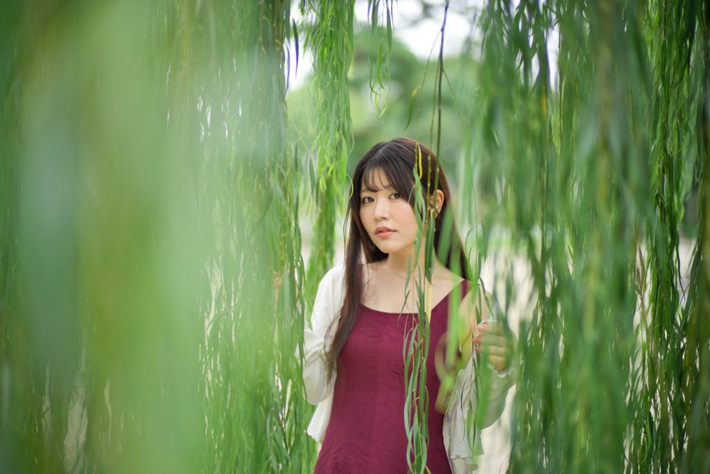 揺れる柳のカーテンの間から佇む桜井こはるさんを捉えたカット(開放F1.0)